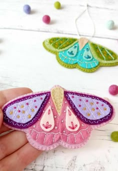 a hand holding a pink and purple moth ornament on a white table with beads