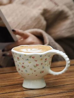 a coffee cup sitting on top of a wooden table next to a person holding a book