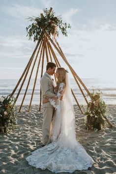 a bride and groom kissing on the beach in front of a teepeet arch