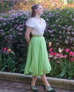 a woman is standing in front of flowers wearing a green skirt and white shirt with pink tulips behind her