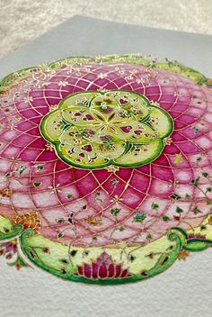 a pink and green circular design on white paper with gold trim around the edges, sitting on a table