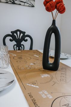 a black vase with three red flowers in it on a cardboard table top next to chairs