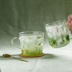 a person holding a glass cup with flowers painted on it and spoon next to it