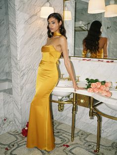 a woman standing in front of a bathroom sink wearing a yellow dress and posing for the camera