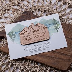 a wooden save the date card on top of a cutting board with mountains in the background