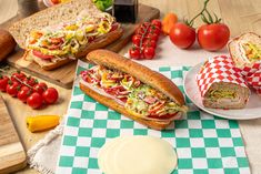 two sub sandwiches sitting on top of a green and white checkered table cloth next to sliced tomatoes