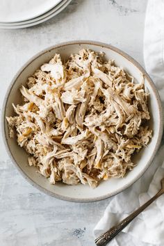 a white bowl filled with shredded chicken next to a fork and napkin on top of a table