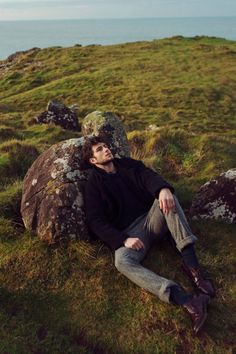 a man laying on top of a lush green field next to a large rock near the ocean