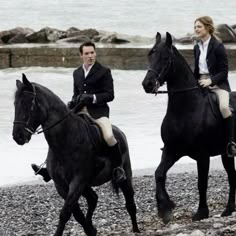 two people riding on the backs of black horses next to water and rocky beach area