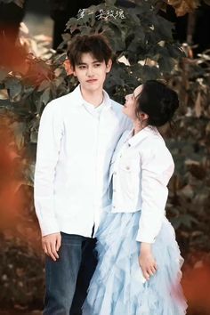 a young man and woman standing next to each other in front of some trees with leaves