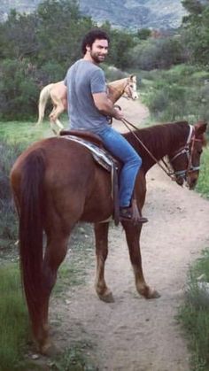 a man riding on the back of a brown horse down a dirt road next to a forest