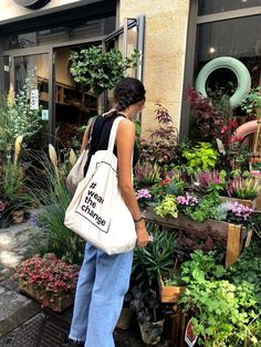 a woman carrying a tote bag in front of a flower shop filled with plants