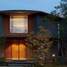 a modern house with wooden doors and windows at night in front of the tree lined driveway