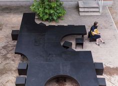 a woman sitting on a bench in front of a black piece of art that looks like a letter