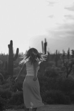 a woman is walking in the desert with her hair blowing in the wind and cacti behind her