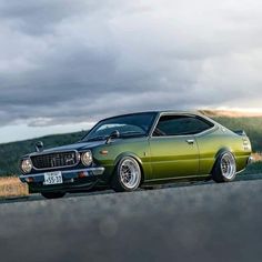 the green car is parked on the side of the road in front of some hills