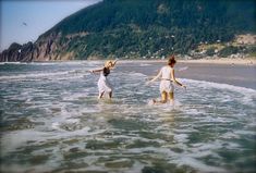 two young women are playing in the water at the edge of the beach, with mountains in the background