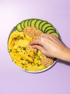 a person is dipping hummus into a bowl with crackers and cucumbers