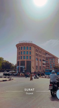 a man riding a motorcycle down a street next to a tall building with the sun in the background