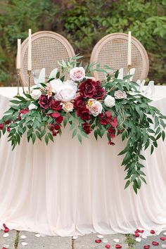 an arrangement of flowers and greenery on a table with two chairs in the background