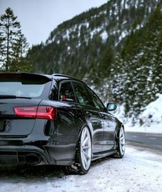 the back end of a black car driving down a snow covered road with mountains in the background