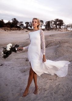 a woman is walking on the beach in a white dress and holding a bridal bouquet