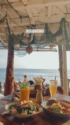 two plates of food sitting on top of a wooden table next to the ocean with people in the background