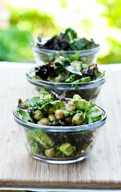 three glass bowls filled with salad on top of a wooden table next to a cutting board