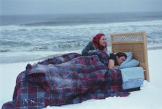 two people laying in bed on the beach with blankets over them and an open suitcase