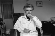 an old man sitting in a chair with his hand on his mouth and looking at the camera