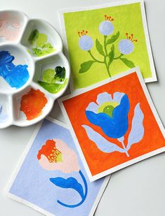 The image shows three square paintings of stylized flowers in bright colors on a white surface. Each painting features a different flower design in contrasting colors: one with blue stems and pink petals on a light blue background, another with a blue flower on an orange background, and the last one with round light blue flowers on a green background. A paint palette with green, blue, and orange paints is visible next to the paintings. Paint Gouache, Flowers Gouache, Modern Watercolor, Guache Paintings Ideas, Guache Paintings Easy, Gouache Illustration, Watercolor Marker Art, Gouache Painting Ideas, Gouache Flowers