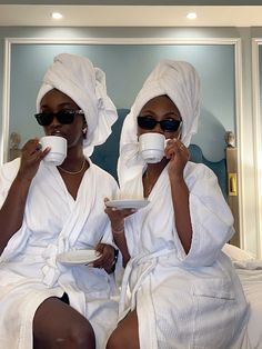 two women in white robes sitting on a bed drinking from cups and saucers with their mouths open