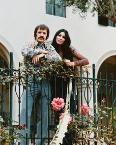 two people standing next to each other on top of a balcony with flowers and plants