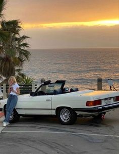 a woman sitting on the hood of a white convertible car by the ocean at sunset