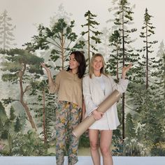 two women standing next to each other in front of a wall with trees on it