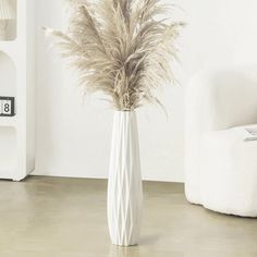 a white vase filled with dry grass on top of a floor next to a chair