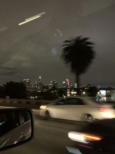 cars driving down the road at night with city lights in the background and palm trees