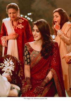 a woman in a red and gold sari sitting on a chair with other people around her