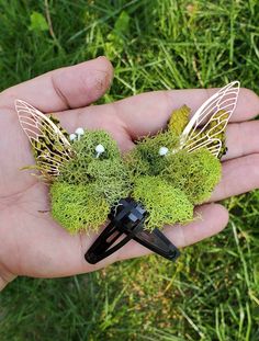 a person holding two hair clips with green moss and white flowers on them in their hand