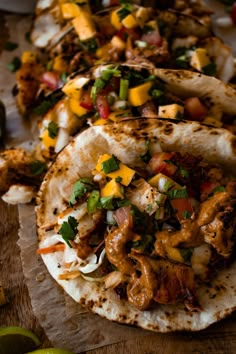 three tacos with meat, cheese and vegetables on paper next to lime wedges