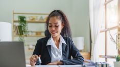 a woman sitting at a desk with a laptop and pen in her hand, writing on paper