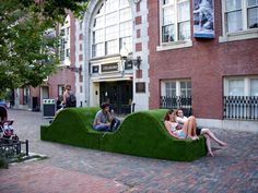 two people sitting on green couches in the middle of a brick sidewalk with buildings behind them