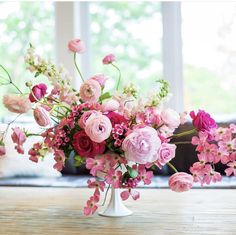 a white vase filled with pink flowers on top of a wooden table next to a window