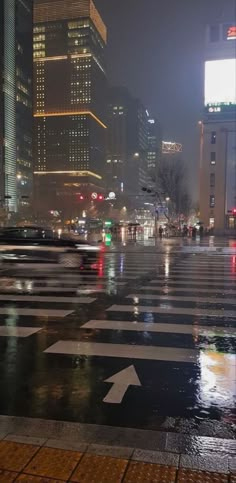 an empty city street in the rain at night with buildings lit up behind it and cars driving on the road