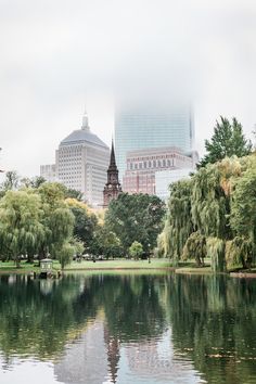 a body of water surrounded by trees and buildings in the background with foggy skies