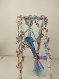 a doll sitting on top of a wooden frame with flowers and leaves around her body