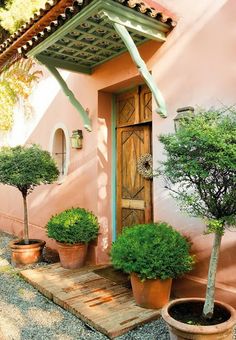 an outdoor area with potted plants and trees in front of a pink building that has a wooden door