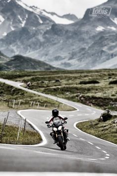 a man riding a motorcycle down a curvy road in front of snow covered mountains