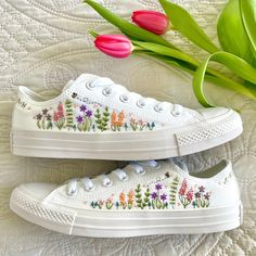 two white shoes with flowers painted on them next to tulips and green leaves