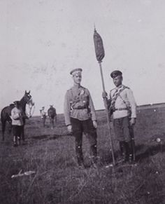 two men standing next to each other in a field with horses and people behind them